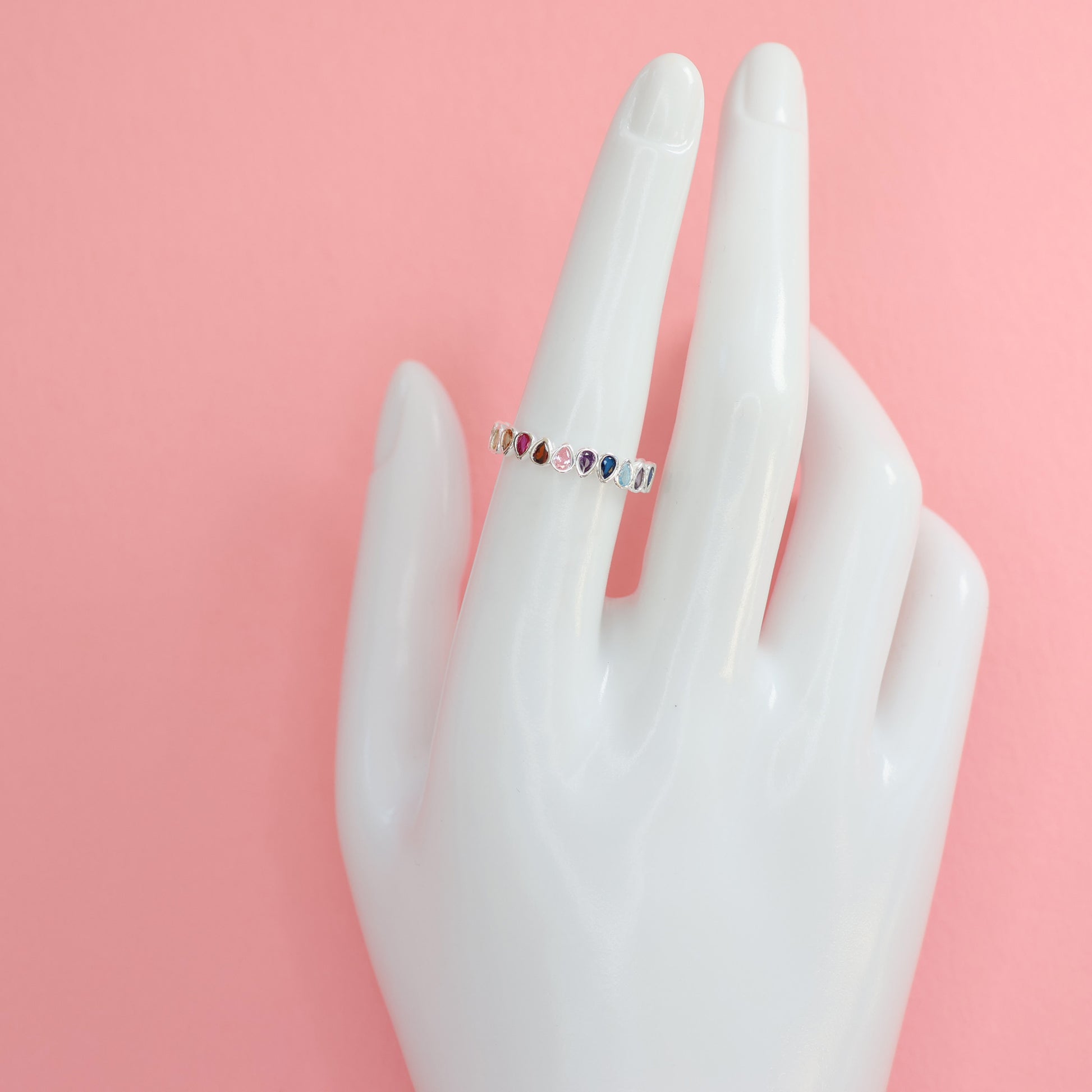 Multi colour rainbow cubic zirconia eternity ring displayed on a mannequin hand against a pink background