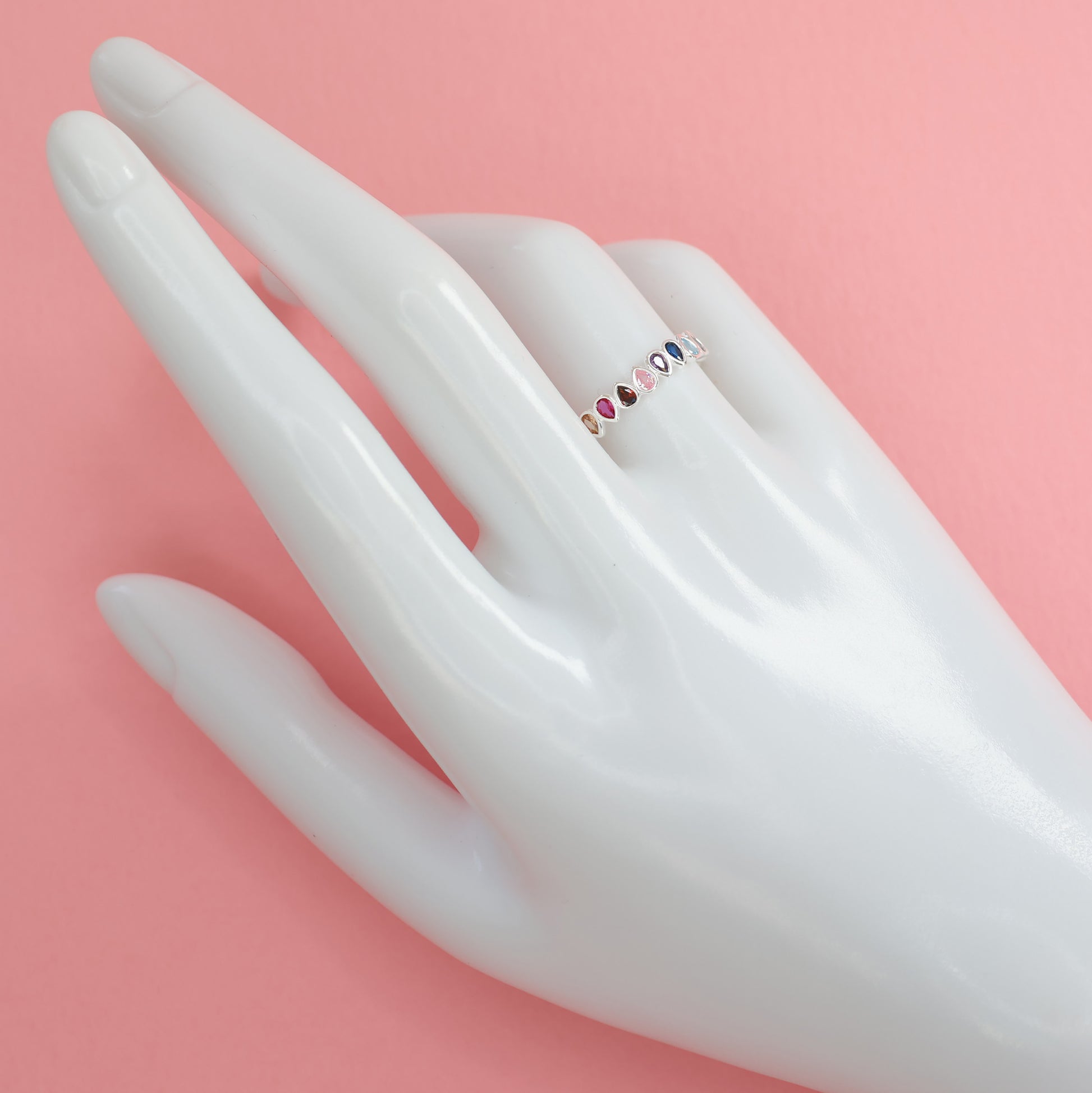 Multi colour rainbow cubic zirconia eternity ring displayed on a hand model against a pink background