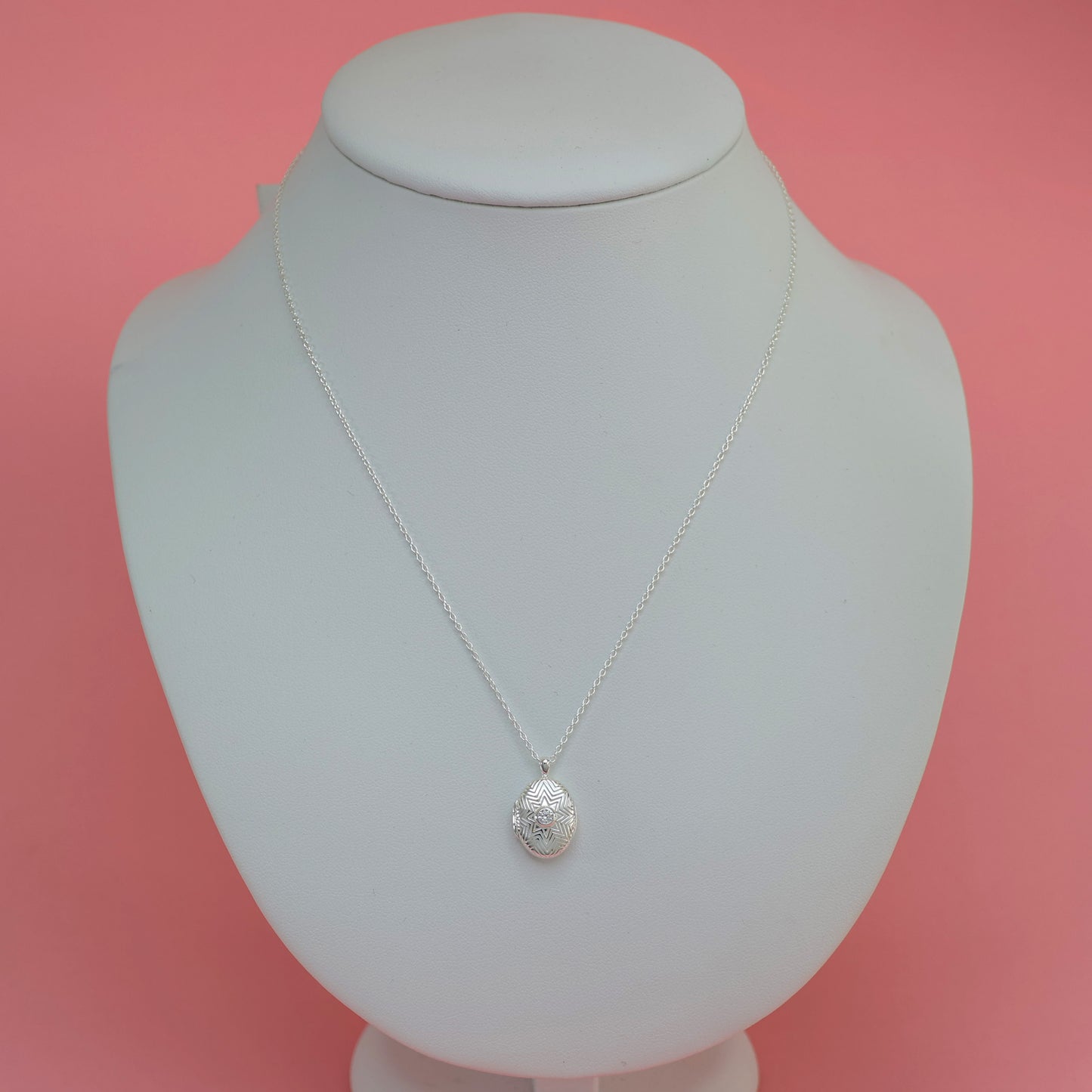 Elegant silver oval locket necklace displayed on a white bust against a pink background